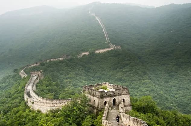 Eine Steinmauer mit Wachtürmen schlängelt sich über weite, üppig grüne Berge unter einem nebligen, bedeckten Himmel, während Menschen darauf entlangwandern.
