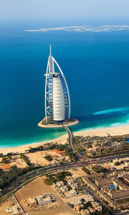 Veduta aerea di un hotel a forma di vela su una piccola isola artificiale, collegata da un ponte a una costa sabbiosa e a una città.