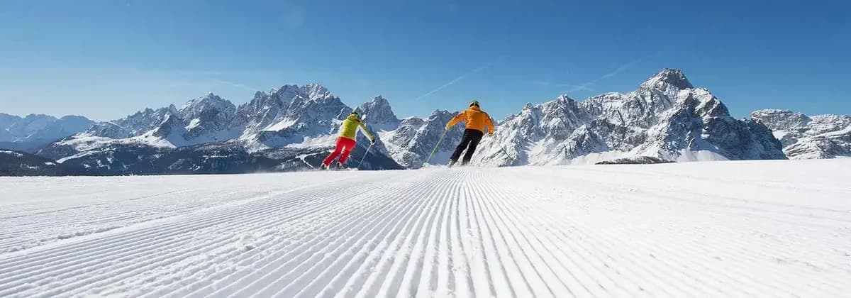 Two skiers descend a freshly groomed snow slope with a panoramic view of a snowy mountain range under a clear sky.