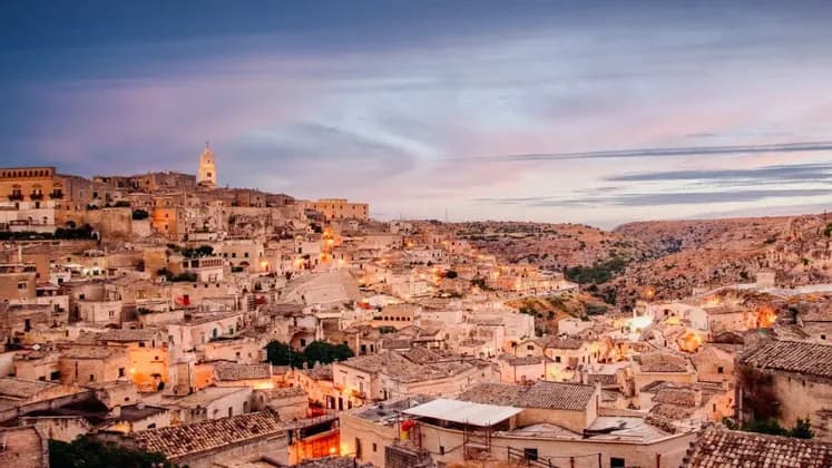 Un borgo di antichi edifici in pietra e un campanile incastonato nella collina, illuminato al crepuscolo accanto a un burrone.