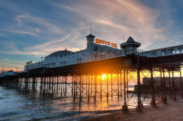Il Molo di Brighton visto dalla spiaggia al tramonto, con luce dorata che splende da dietro e si riflette sul mare.