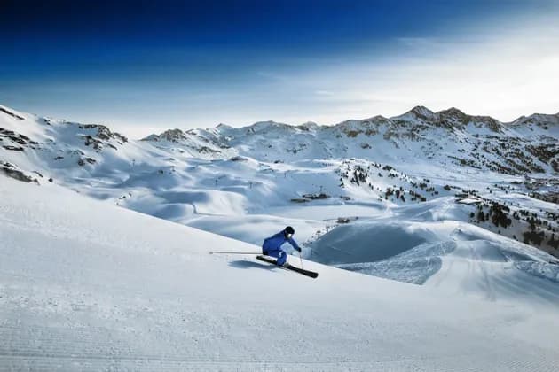 Uno sciatore in tuta blu scende un vasto pendio montano innevato sotto un cielo azzurro limpido.