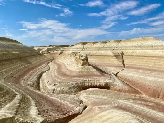 Colorful, layered rock formations of red and white sediment form rolling hills in a canyon under a blue sky.