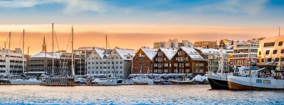 Ein Panorama eines verschneiten Hafens mit Booten, die an einer von traditionellen Gebäuden gesäumten Uferpromenade liegen, bei Sonnenuntergang.
