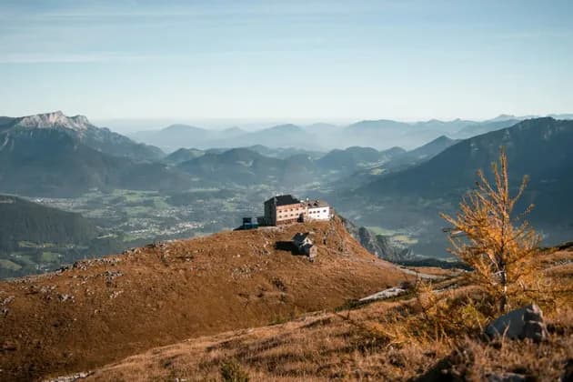 Ein großes Gebäude thront auf einem grasbewachsenen Berggipfel und überblickt ein weites Tal sowie geschichtete Bergketten unter einem klaren Himmel.