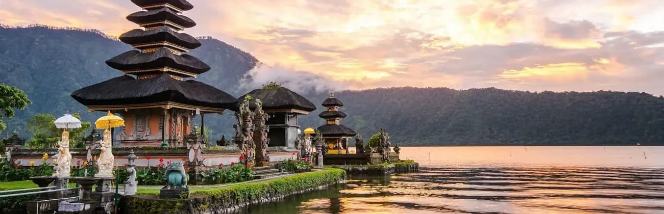Un ornamentado complejo de templos con tejados escalonados se encuentra a orillas de un lago al atardecer, con montañas elevándose en el fondo.