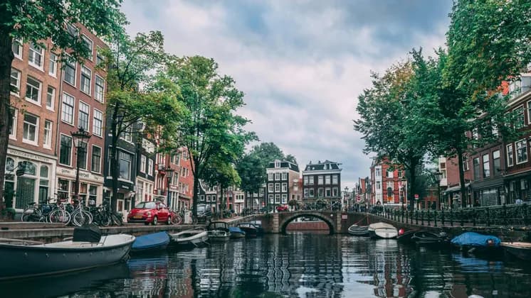 Une vue sur un canal urbain bordé de bâtiments traditionnels, d'arbres verts et de bateaux amarrés, avec un pont de pierre au loin.
