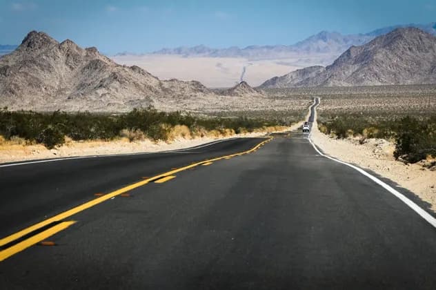 Una carretera pavimentada con líneas amarillas se extiende por un valle desértico, con montañas rocosas visibles a lo lejos bajo un cielo azul.