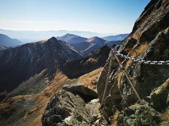 Eine metallene Sicherheitskette verläuft entlang eines schmalen, felsigen Bergpfades mit Blick auf eine weite Bergkette unter klarem Himmel.