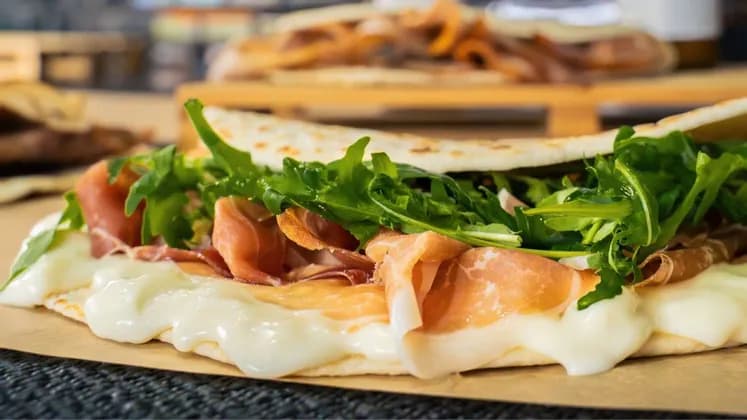 Primo piano di una piadina piegata ripiena di prosciutto, formaggio fuso e rucola fresca su carta marrone.