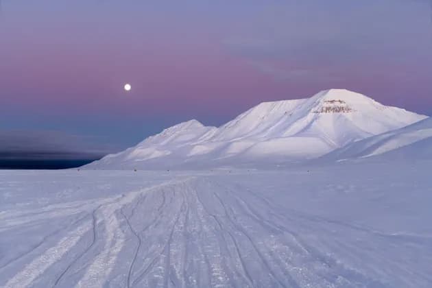 Una luna piena brilla in un cielo crepuscolare violaceo sopra una vasta catena montuosa innevata con tracce nella neve.