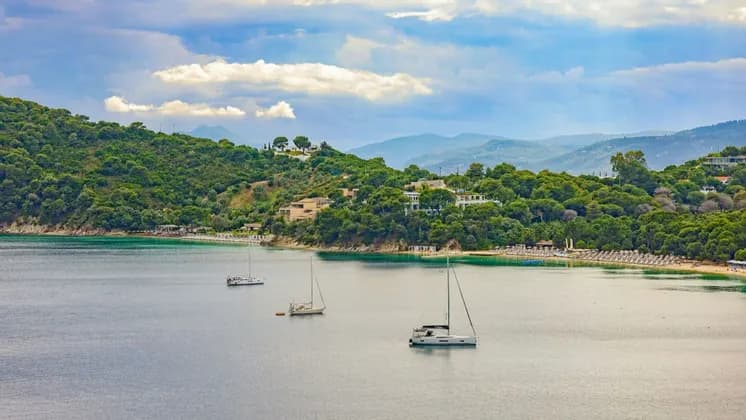 Tre barche a vela galleggiano in una baia tranquilla accanto a una costa verde e collinare con edifici sparsi sotto un cielo nuvoloso.