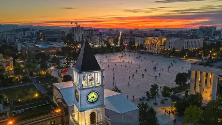 Una vista aerea di un'ampia piazza cittadina con una torre dell'orologio in primo piano e persone che passeggiano durante un tramonto colorato.