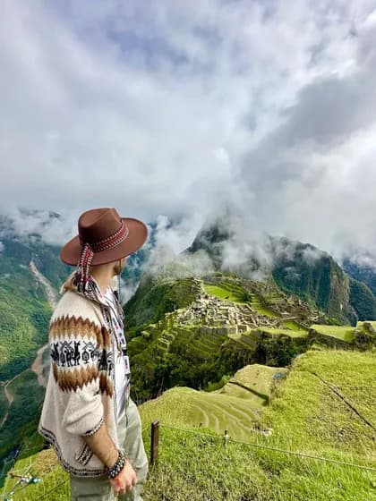 Un uomo con un cappello e un maglione a fantasia osserva antiche rovine costruite su verdi montagne terrazzate sotto un cielo nuvoloso.