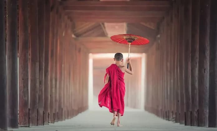 Un giovane monaco a piedi nudi in tuniche rosse cammina lungo un corridoio di legno tenendo un ombrello rosso.