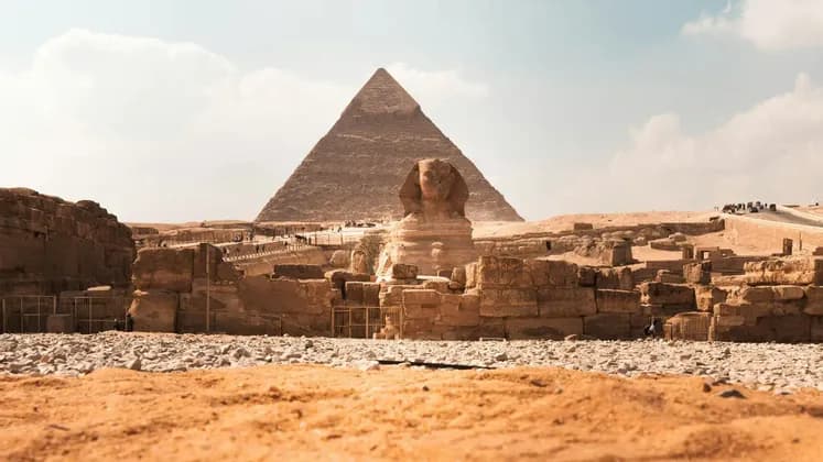Una vista desde el suelo de la Gran Esfinge con una gran pirámide al fondo, rodeada de antiguas ruinas de piedra bajo un cielo brillante.