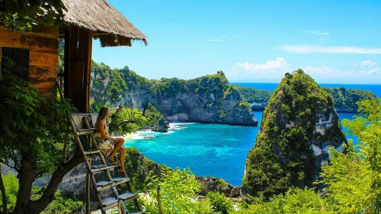 Una mujer con un vestido floral se sienta en una escalera de madera junto a una casa en el árbol, con vistas a una bahía tropical con acantilados y agua turquesa.