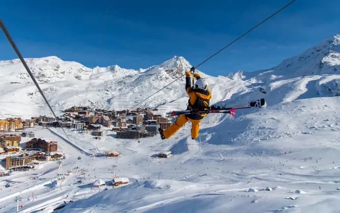 Une personne en équipement de ski descend une tyrolienne avec des skis sur le dos, au-dessus d'un village de montagne enneigé.