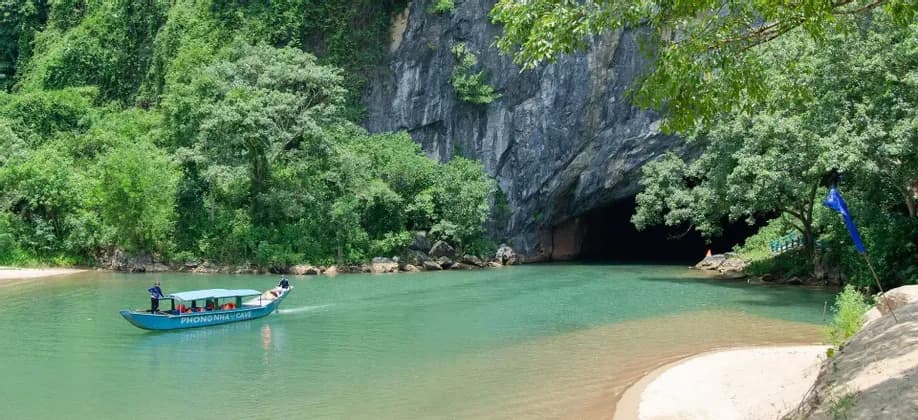 Una barca turistica blu con persone naviga su un fiume turchese, dirigendosi verso una grande apertura di grotta alla base di una montagna boscosa.