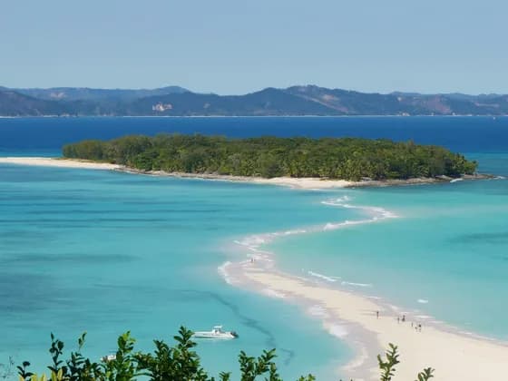 Eine sich schlängelnde weiße Sandbank verbindet sich mit einer kleinen, grünen Insel in türkisfarbenem Wasser, mit fernen Bergen im Hintergrund.
