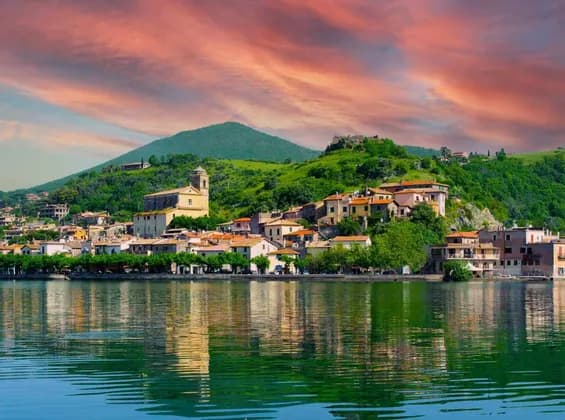 Un borgo lacustre con una chiesa e case dai tetti in terracotta riflesse nell'acqua sotto un cielo al tramonto dai colori vivaci.