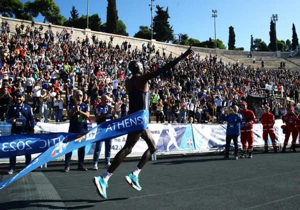 Un corridore taglia il traguardo della maratona di Atene, rompendo il nastro blu mentre la folla esulta dagli spalti dello stadio.