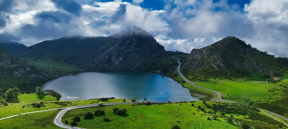 Una vista aerea di una strada tortuosa lungo un lago calmo, incastonata tra verdi colline e montagne sotto un cielo nuvoloso.