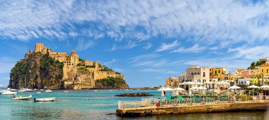 Un castello si erge su una scogliera rocciosa che domina una città costiera e una baia piena di barche sotto un cielo azzurro.