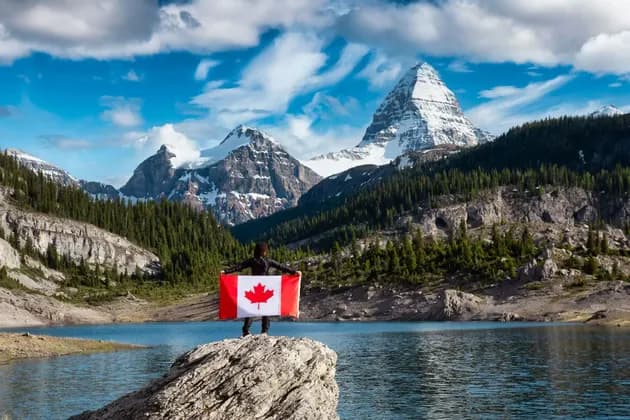 Eine Person hält eine große kanadische Flagge auf einem Felsen mit Blick auf einen Alpensee und schneebedeckte Berge.