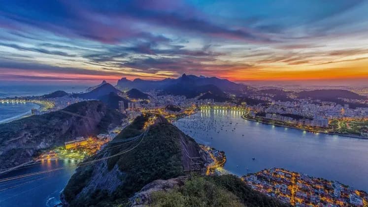 A panoramic view of an illuminated coastal city nestled between mountains and a bay under a colorful sunset sky.