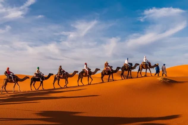 A WeRoad group trip rides a line of camels over orange desert sand dunes under a partly cloudy sky.