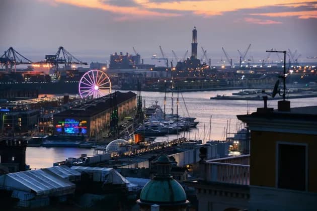 Una vista aerea di un porto al crepuscolo, che mostra una ruota panoramica illuminata, un faro, barche ormeggiate e gru industriali.