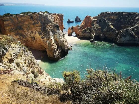 A secluded cove with a natural rock arch is surrounded by golden cliffs and turquoise water, viewed from above on a sunny day.