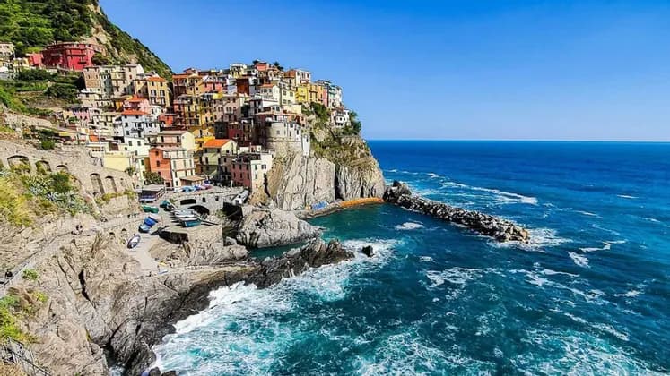 Una vista soleggiata di un colorato villaggio su una scogliera rocciosa che si affaccia su un mare blu profondo con una diga foranea in pietra.