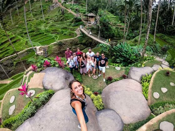 Una mujer se toma una selfie desde un ángulo alto con un grupo de WeRoad posando en una ladera verde aterrazada con grandes rocas.