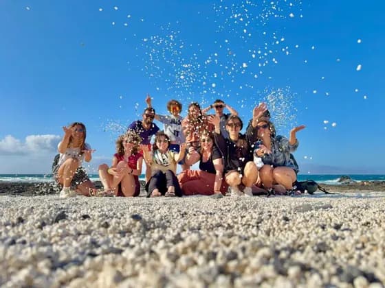 Un viaggio di gruppo WeRoad lancia sassolini bianchi in aria mentre posa per una foto su una spiaggia assolata.