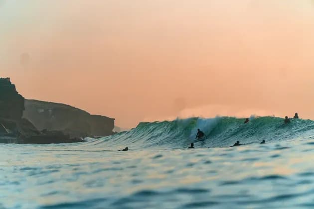 Viaggio di gruppo WeRoad di surfisti nell'oceano, con un surfista su una grande onda verde vicino a scogliere al tramonto.