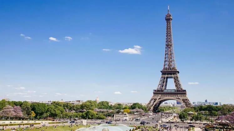 Una vista diurna amplia de la Torre Eiffel, con un primer plano de árboles verdes, un parque y el horizonte de la ciudad bajo un cielo azul claro.