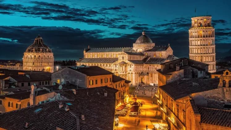 Veduta dall'alto di Torre Pendente, cattedrale e battistero illuminati al crepuscolo, sopra i tetti di una città storica.