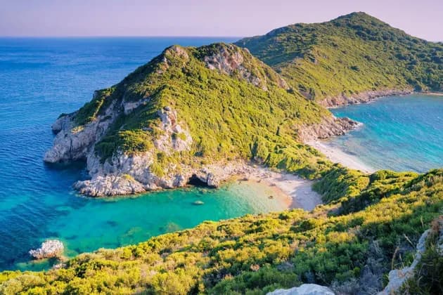 A lush green, rocky headland forms a small cove with a sandy beach and clear, turquoise water on a sunny day.