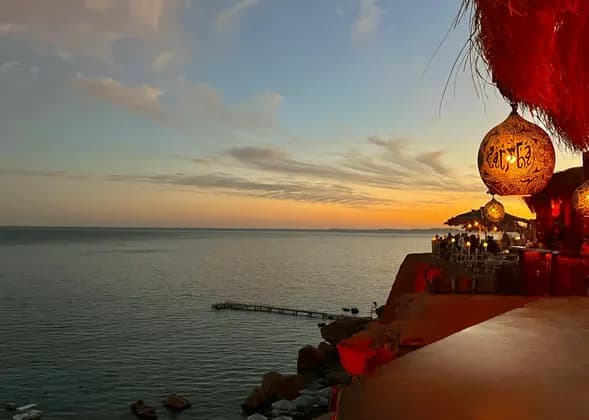 Vista da un ristorante sulla scogliera con affaccio sul mare durante un tramonto colorato, con una lanterna ornata in primo piano.