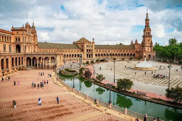 Una vista elevada de una gran plaza con un edificio semicircular ornamentado, un canal con puentes, una fuente central y gente caminando alrededor.