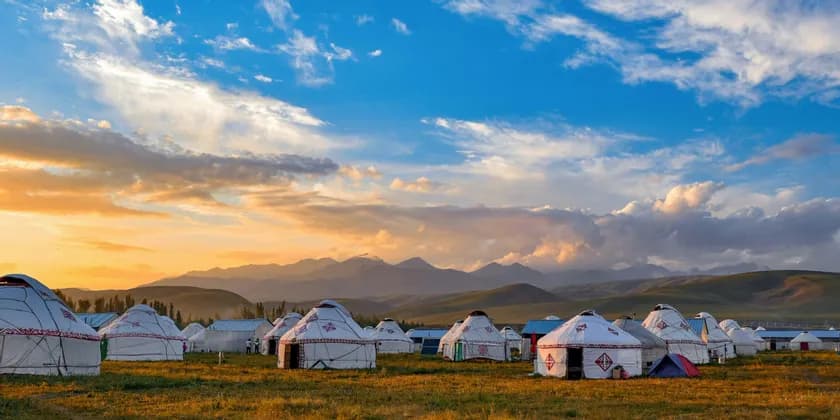A camp of traditional white yurts in a grassy field with distant mountains under a colorful sunset sky.
