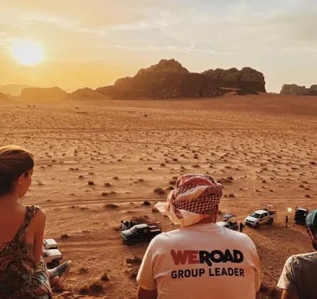 Un viaggio di gruppo WeRoad ammira il tramonto su un vasto paesaggio desertico con formazioni rocciose distanti e diverse jeep.