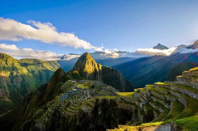 A panoramic view of ancient stone ruins and terraces on a green mountainside with a prominent peak lit by sunlight.