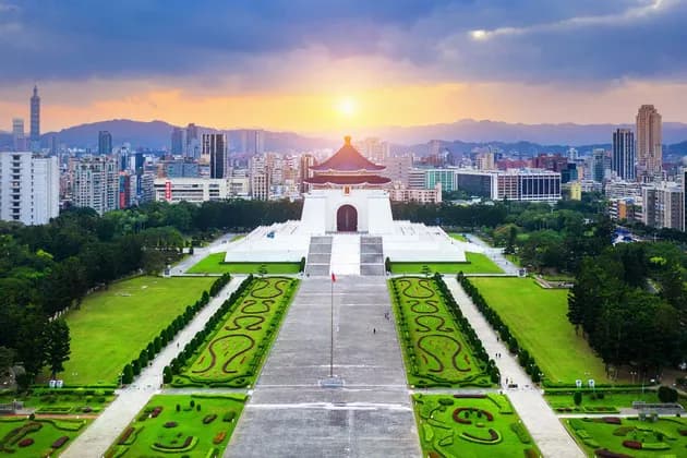 Una veduta aerea di una sala commemorativa bianca con un tetto blu, sullo sfondo di uno skyline moderno e montagne al tramonto.