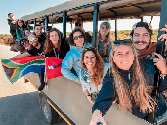Un gruppo WeRoad sorridente posa per un selfie all'interno di un veicolo safari aperto, mostrando una bandiera sudafricana.