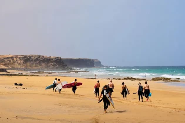 Un groupe WeRoad en combinaisons de surf, planches à la main, marche sur une plage de sable vers l'océan, avec des falaises au loin.