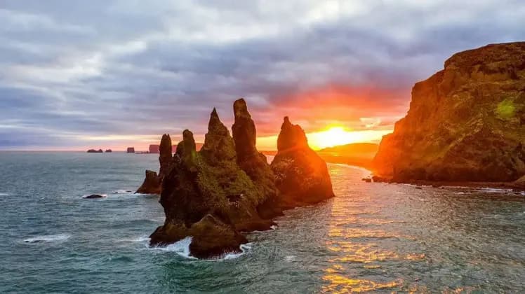 Zackige, moosbewachsene Felsnadeln ragen aus dem Meer, während die Sonne untergeht und ein orangefarbenes Glühen auf das Wasser wirft.
