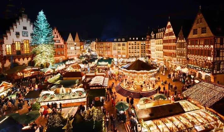 An elevated view of a bustling Christmas market at night, with a large illuminated tree, a glowing carousel, and market stalls.